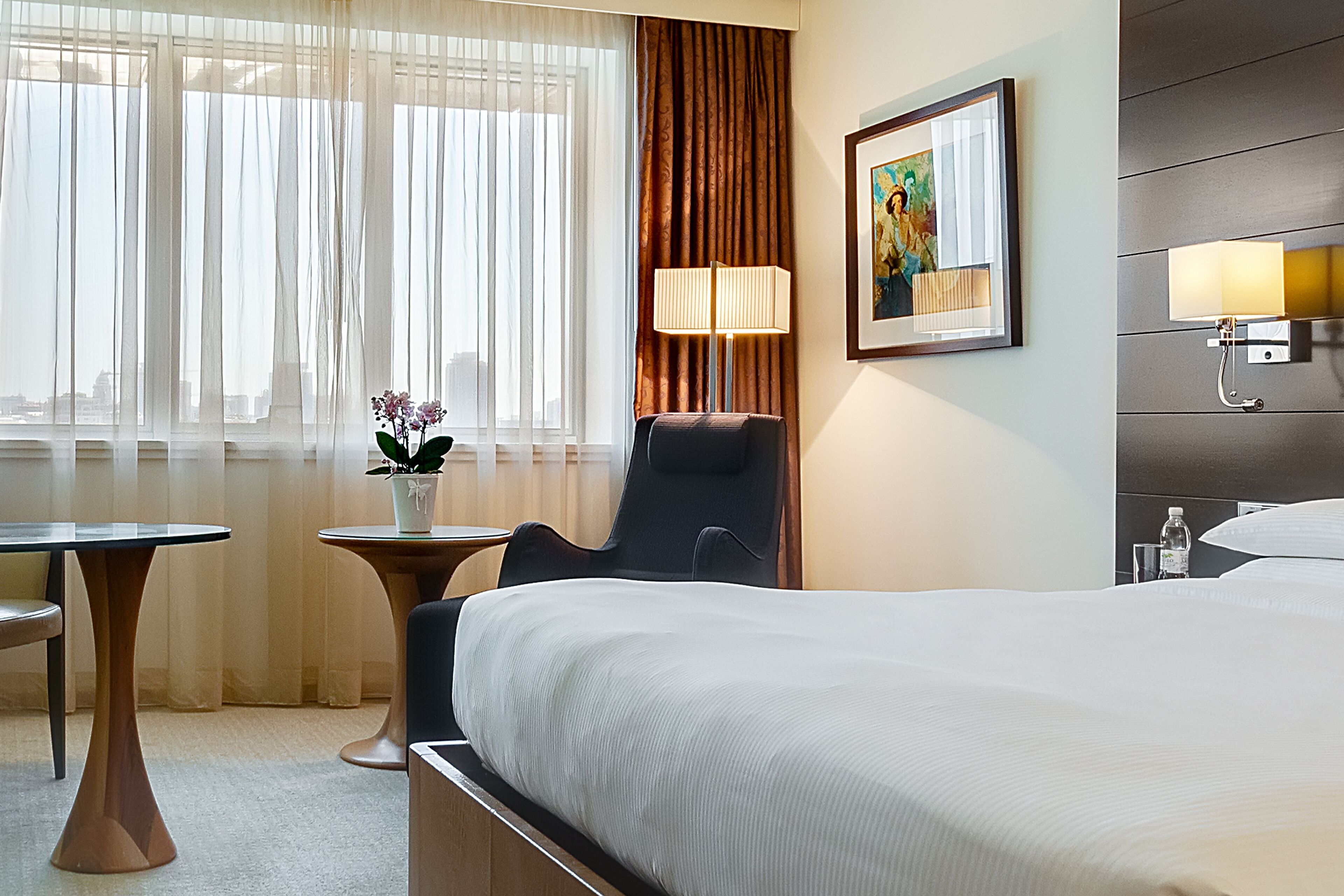 A hotel room with a bed, a desk, a chair, a lamp, and a painting on the wall.