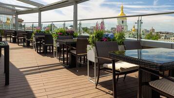 Una terraza al aire libre con mobiliario de mimbre, plantas en macetas y vista a un paisaje urbano con cúpulas doradas.