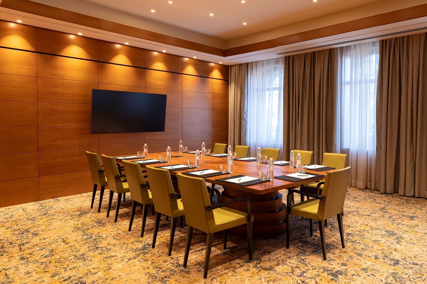 A conference room with a large table, chairs, a flat-screen TV, and a window with curtains.