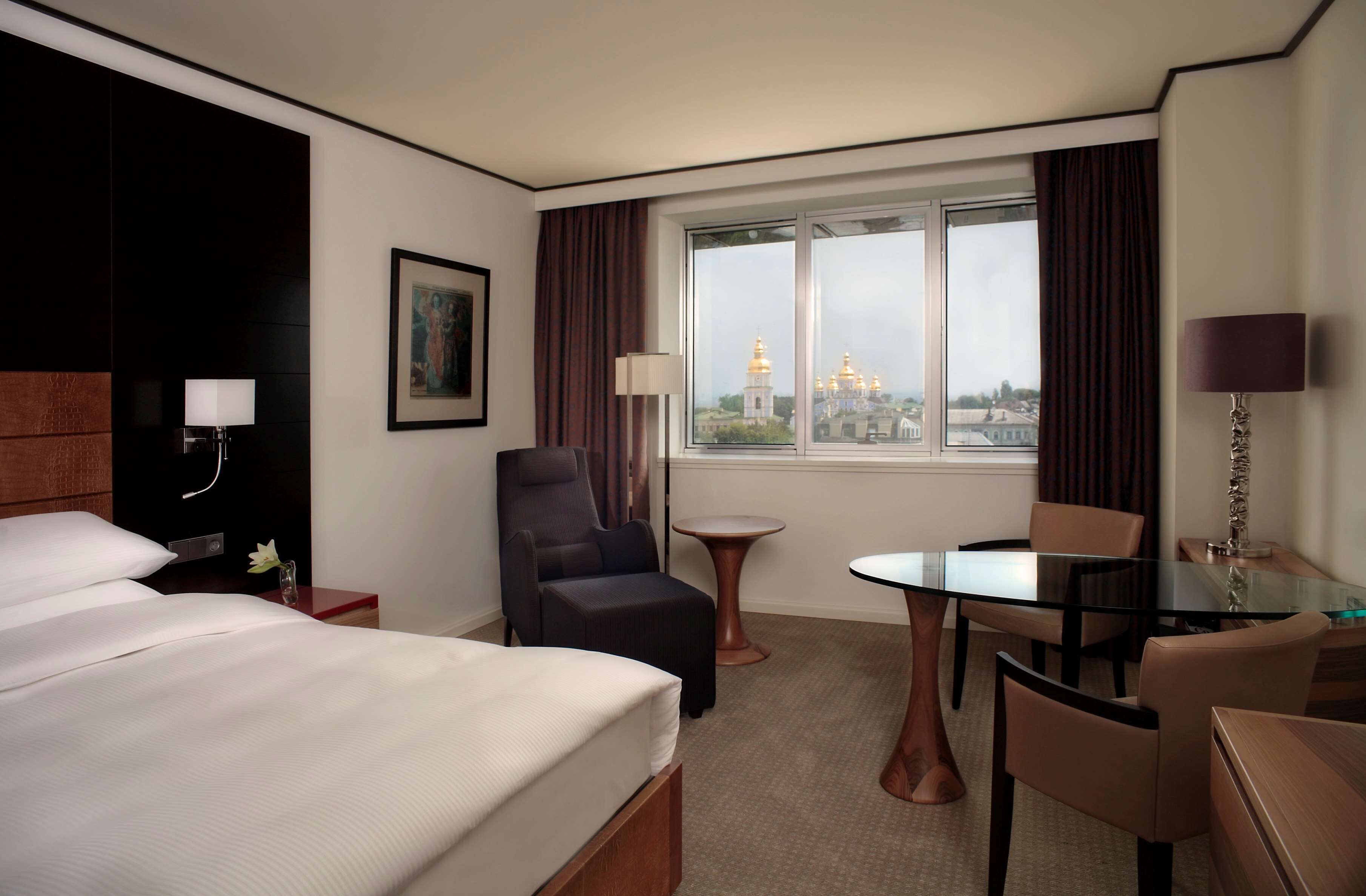 A hotel room with a large bed, a sitting area with a table and chairs, a small round table, a lamp, and a view of a building with golden domes.