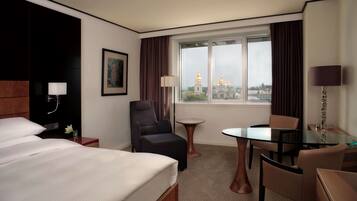 A hotel room with a large bed, a sitting area with a table and chairs, a small round table, a lamp, and a view of a building with golden domes.