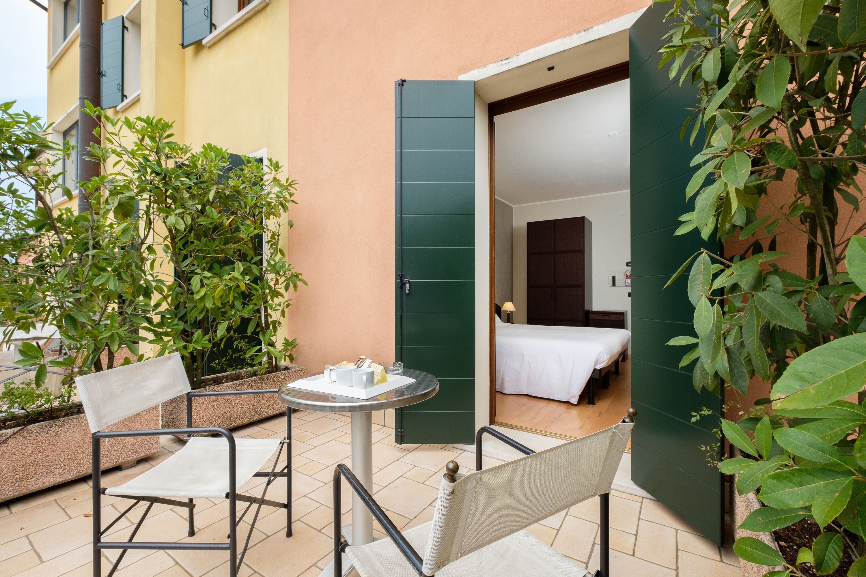 A small outdoor seating area with two chairs and a table, a door leading to a bedroom, and greenery around.