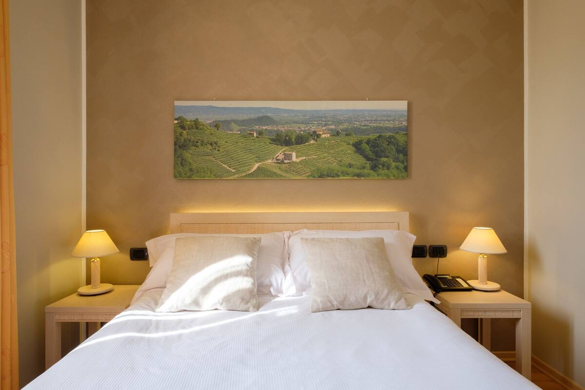 Habitación de hotel con una cama grande, dos mesitas de noche con lámparas y un cuadro de paisaje en la pared.