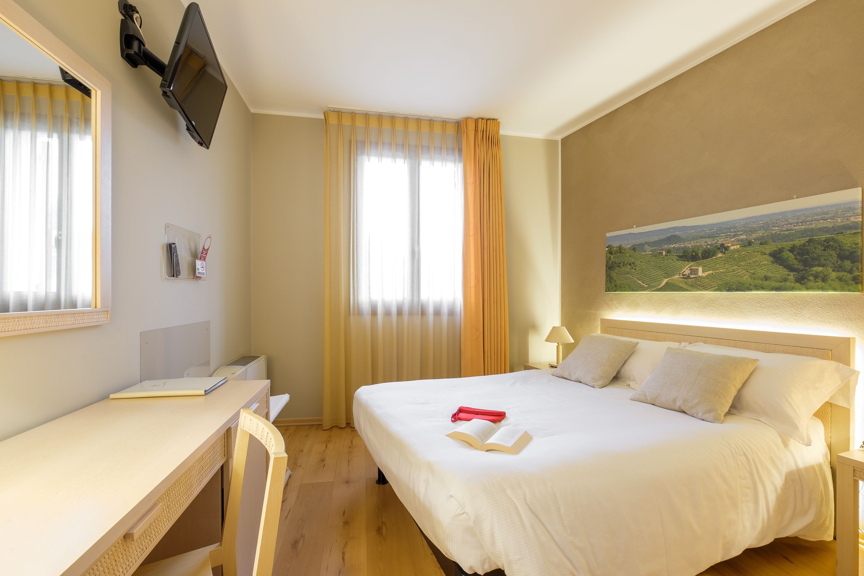 A hotel room with a bed, a desk, a TV mounted on the wall, and a painting on the wall.