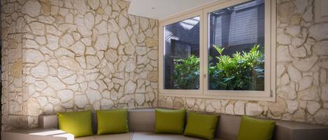 A curved sofa with green cushions against a stone wall, a window with a view of greenery, and a ceiling with recessed lighting.