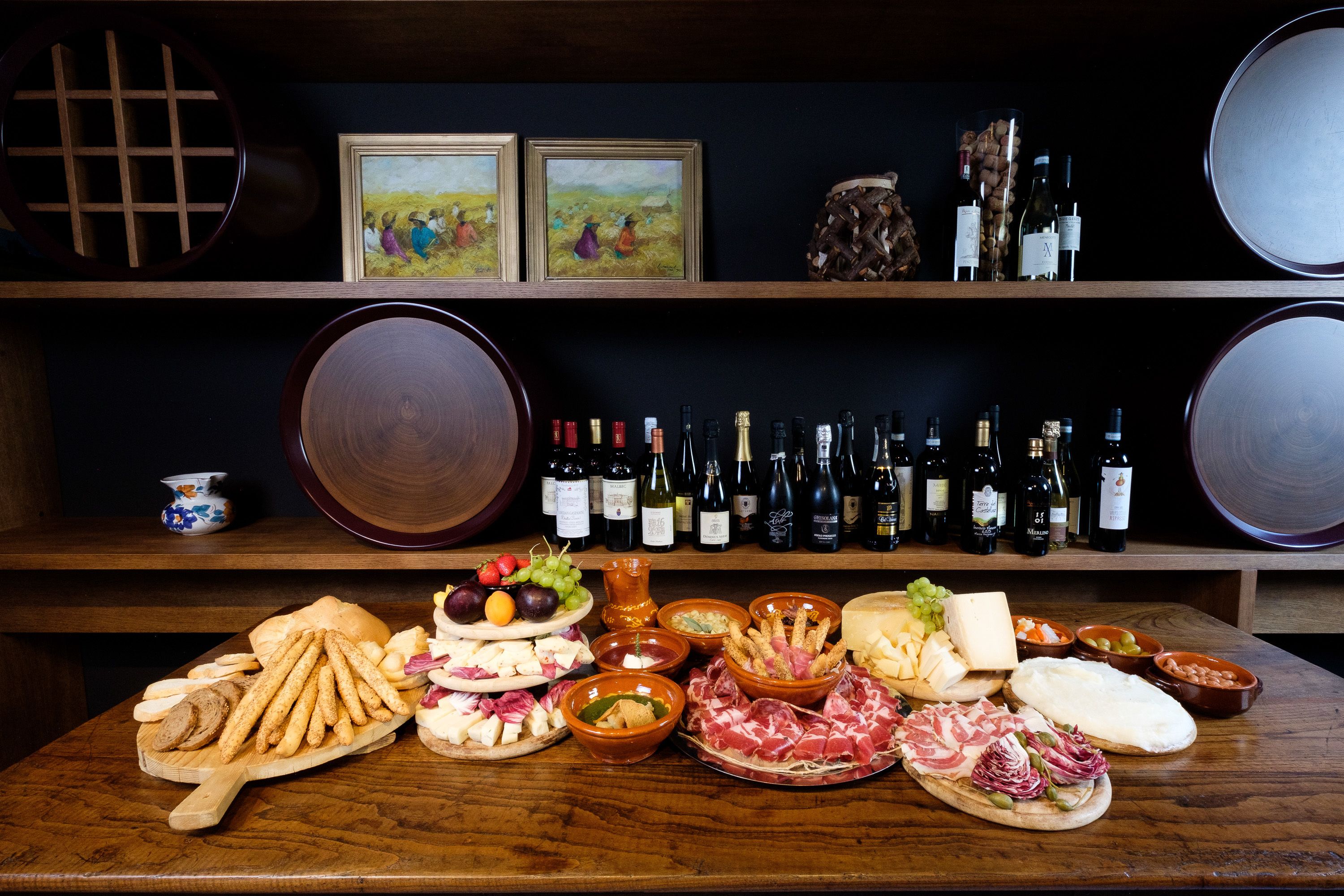 A wooden table with various cheeses, meats, and breads, and a wine rack with bottles behind it.