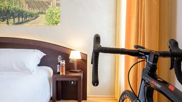 Quarto de hotel com uma cama, uma mesinha de cabeceira com abajur, uma garrafa e uma bicicleta.