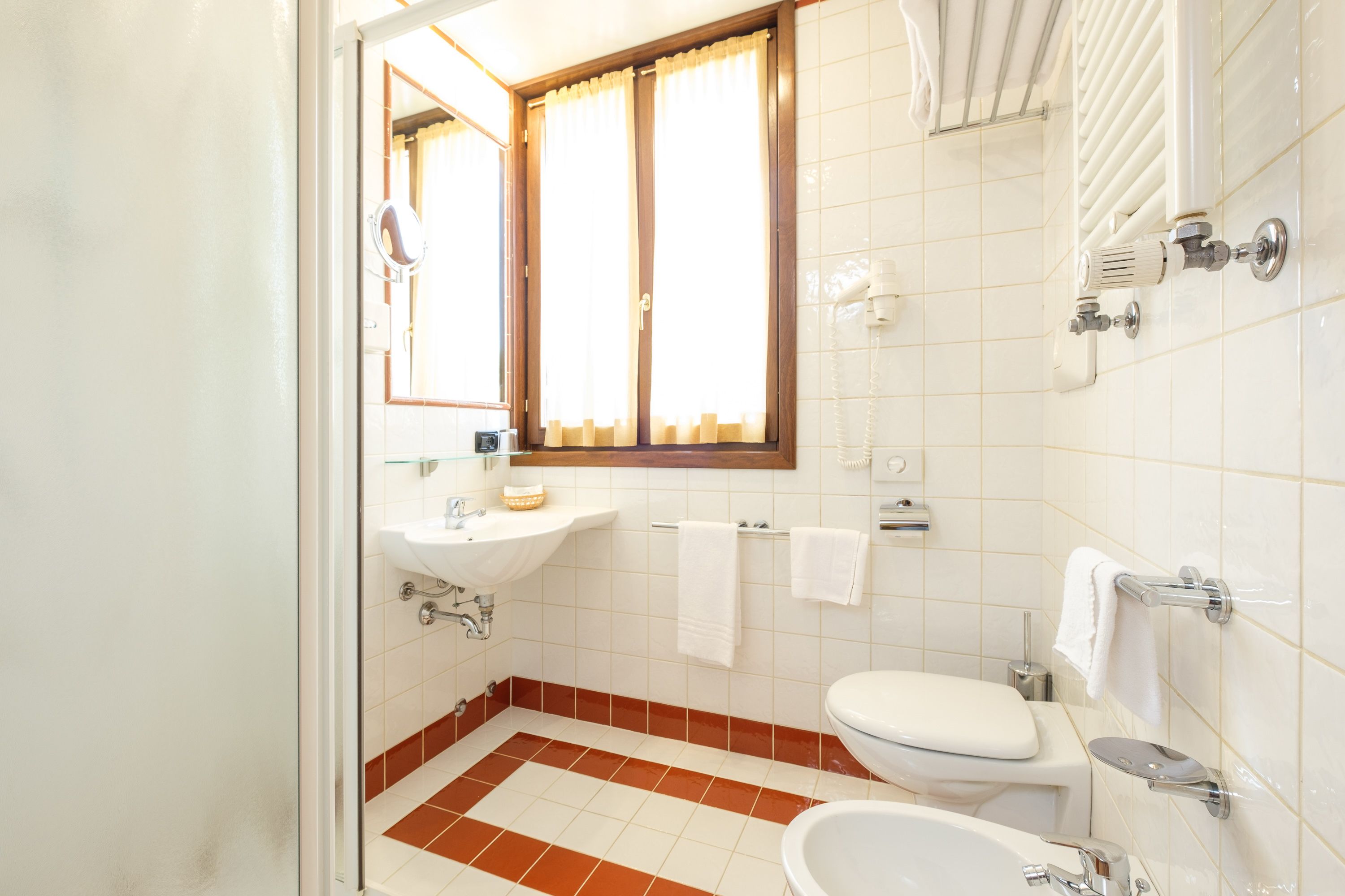 A bathroom with a sink, toilet, and bidet, tiled in white and brown, with a window and a mirror.