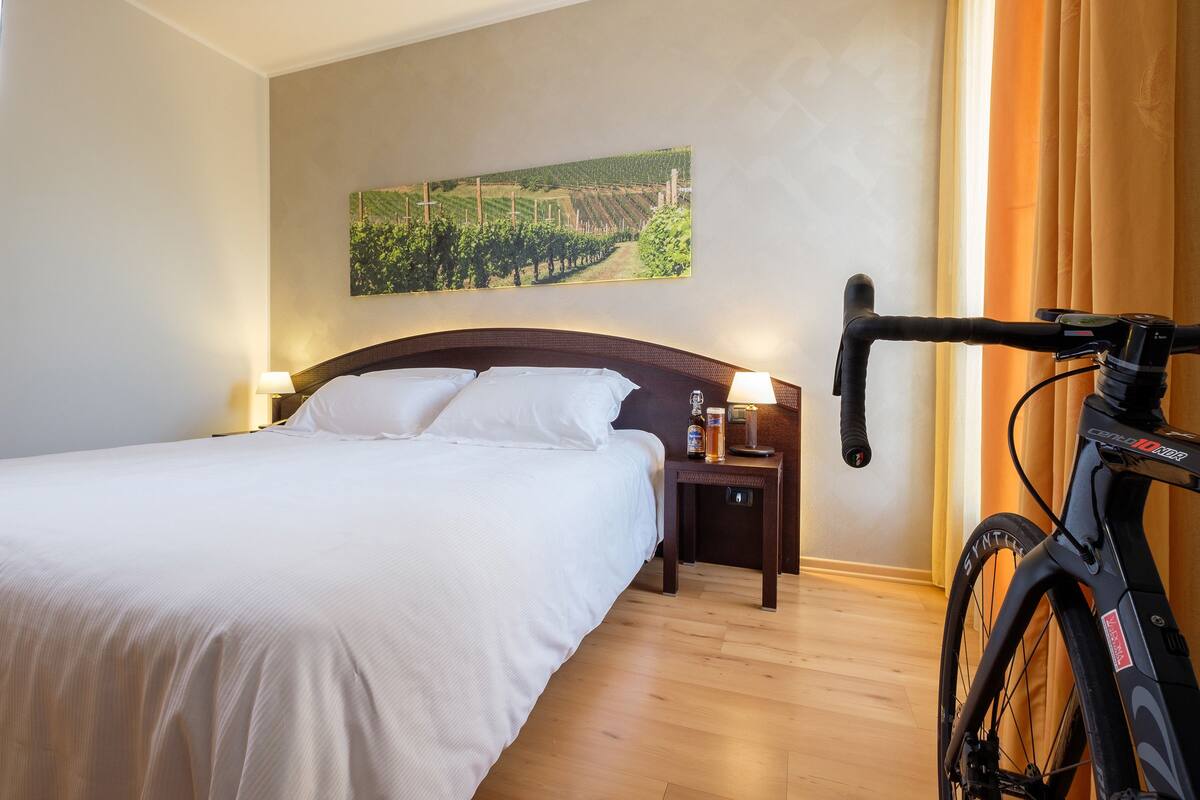 Una habitación de hotel con una cama, una mesita de noche, una bicicleta y un cuadro de un viñedo en la pared.