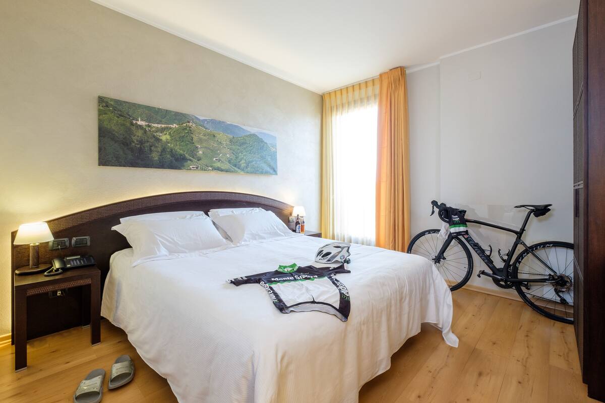 Habitación de hotel con cama, mesita de noche con lámpara, bicicleta y un cuadro en la pared.