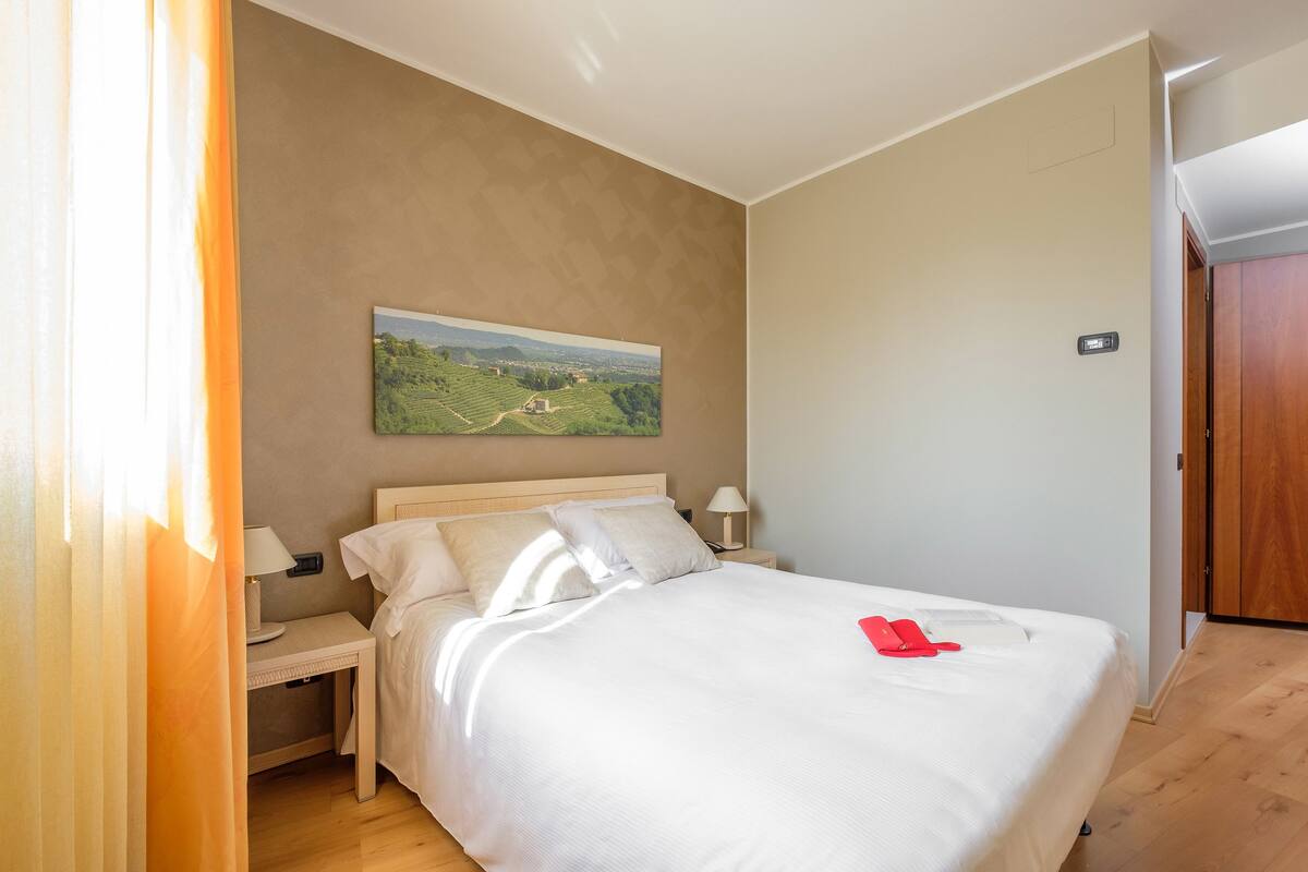 Una cama bien tendida con sábanas blancas, una mesita de noche con una lámpara, un cuadro de un paisaje en la pared y una puerta de madera a la derecha.