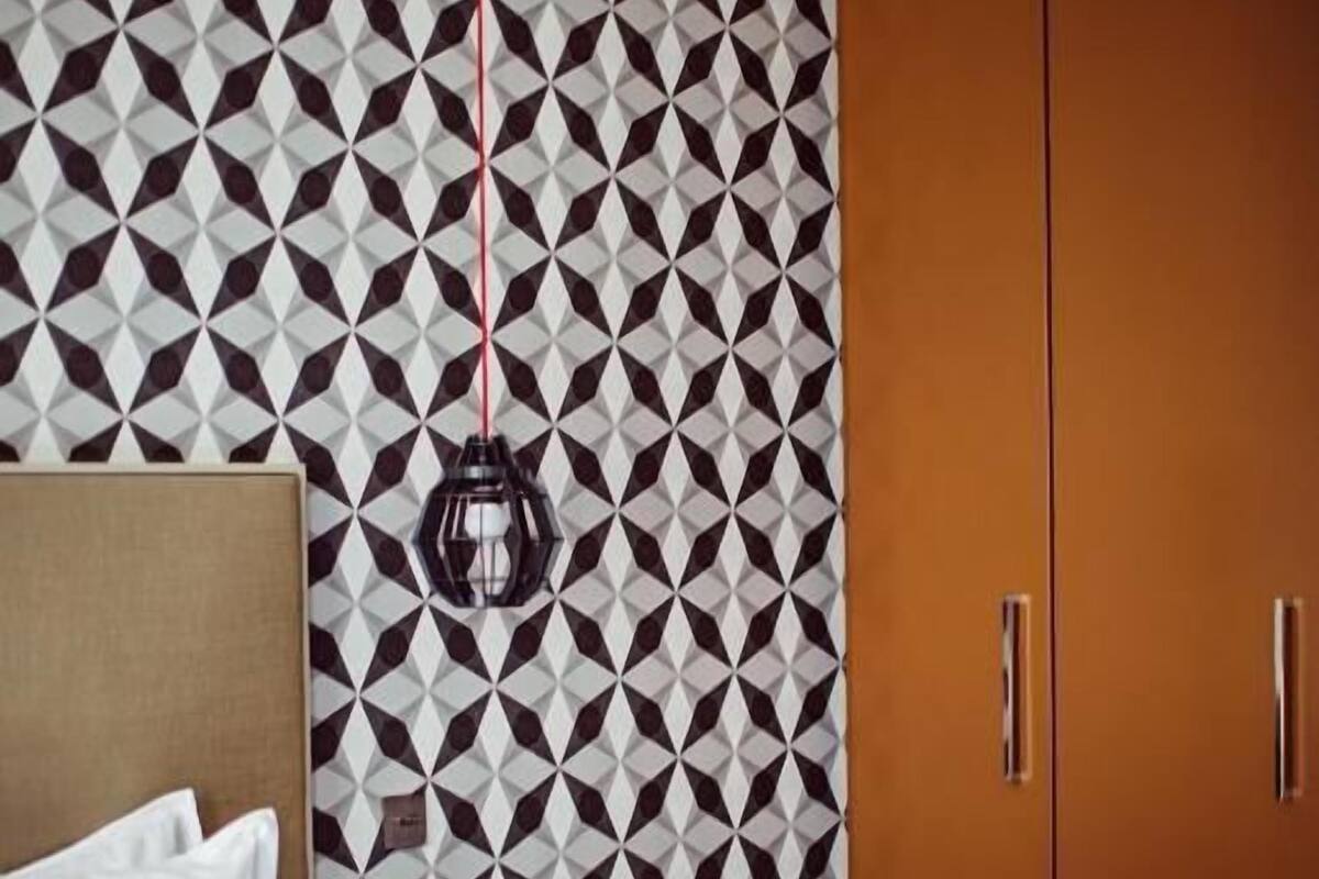 A hotel room with a geometric patterned wall, a bed with white bedding, a bedside table with a book and a lamp, and a wardrobe with two doors.