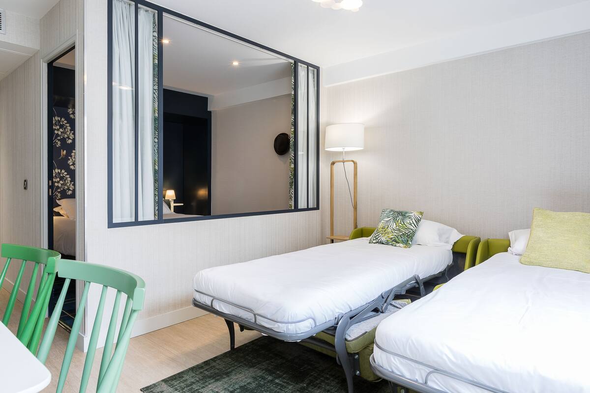 A hotel room with two single beds, a green chair, a small table, and a mirror.
