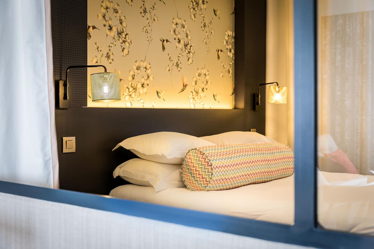 A hotel room with a bed, pillows, a wall-mounted light fixture, and a floral wallpaper design.