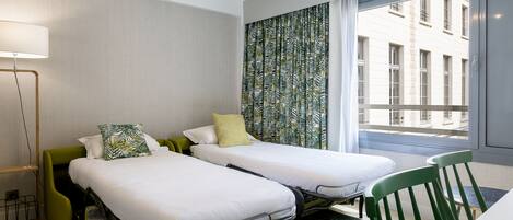Une chambre d’hôtel avec deux lits simples, un fauteuil vert, un bureau et une grande fenêtre donnant sur des immeubles.
