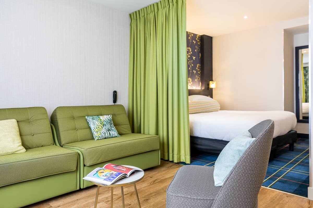 A hotel room with a green sofa, a grey armchair, a small table with a magazine, a bed with a white comforter, and a green curtain.