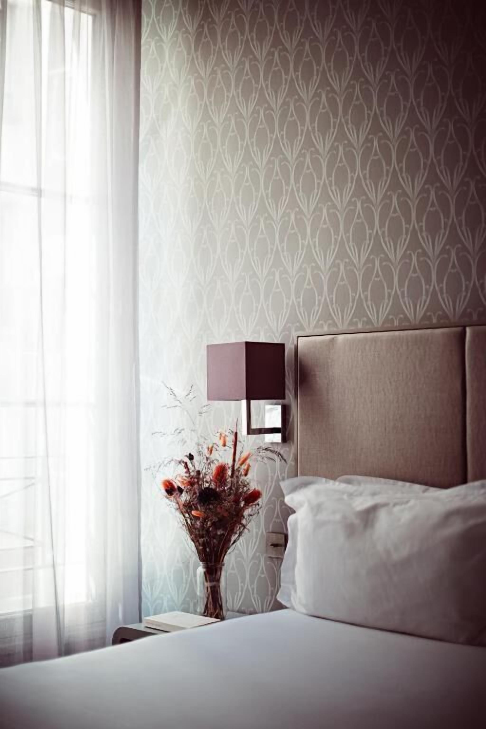 A hotel room with a bed, a nightstand, a vase with dried flowers, a wall-mounted lamp, and a window with sheer curtains.