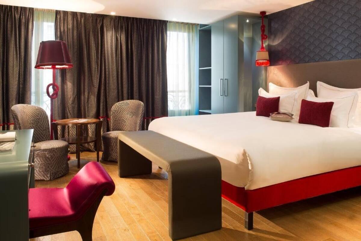 A hotel room with a large bed, a desk, a chair, a bench, and a lamp.