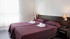 Hotellihuone, jossa on kaksi sänkyä, ja jokaisessa on violetti sängynpeite. Lisäksi huoneessa on yöpöytä, jossa on lamppu, ja pieni ruukkukasvi.