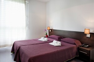 Quarto de hotel com duas camas, cada uma com colcha roxa, mesa de cabeceira com abajur e um pequeno vaso de plantas.