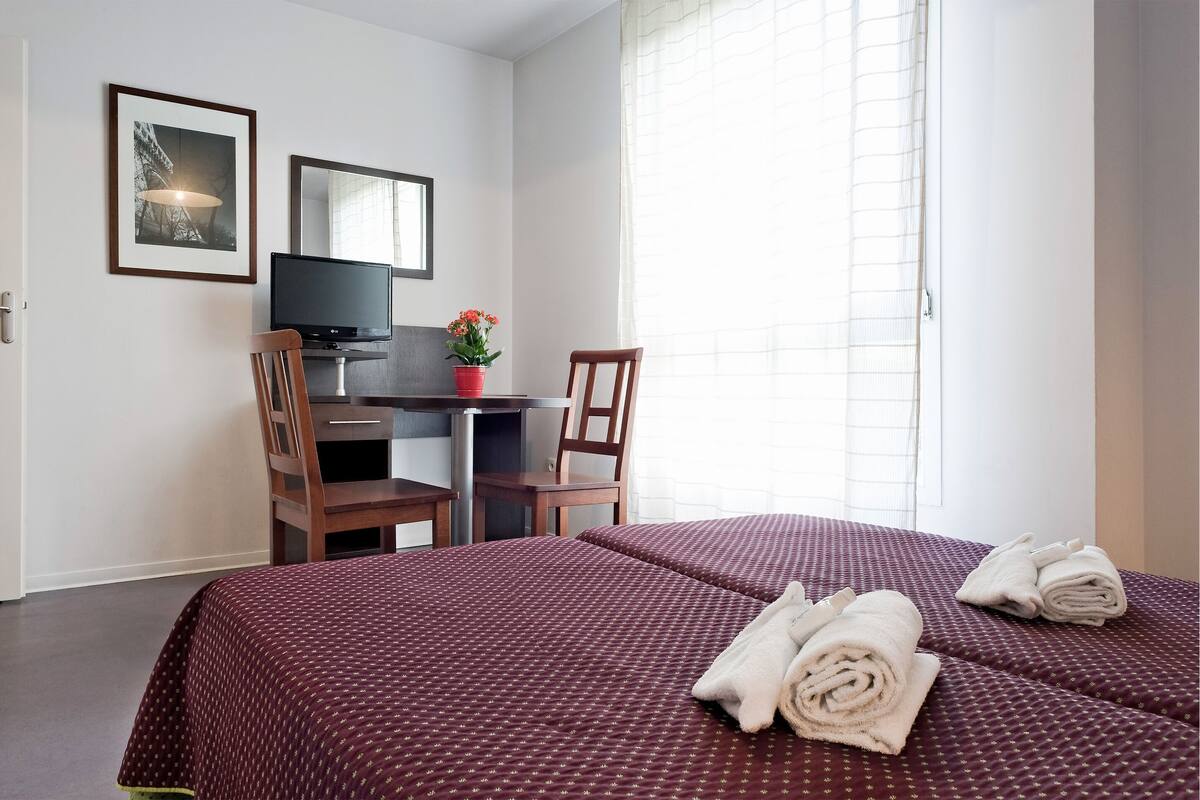 A hotel room with two beds, a desk, a chair, a TV, and a window with curtains.