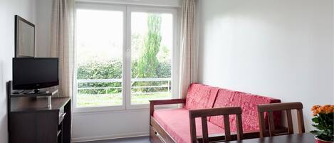 Ein Zimmer mit einem Bett, einem Fernseher, einem Tisch und einem Fenster mit Vorhängen.