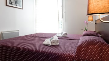 Habitación de hotel con dos camas, una ventana, un radiador, una lámpara fijada a la pared y un cuadro enmarcado.