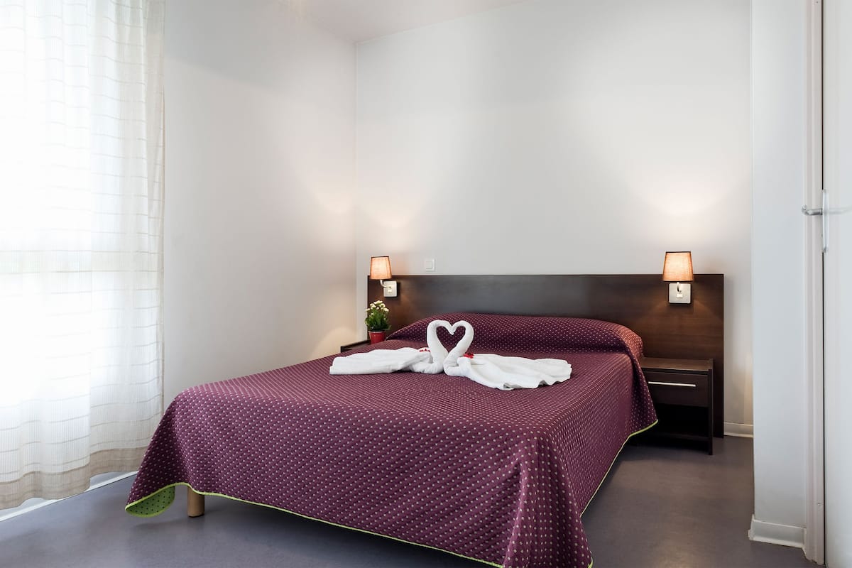 A neatly made bed with a heart-shaped towel arrangement, a bedside lamp, and a small potted plant.