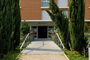 Entrada de hotel com balcão de recepção, passarela coberta e árvores altas de ambos os lados.