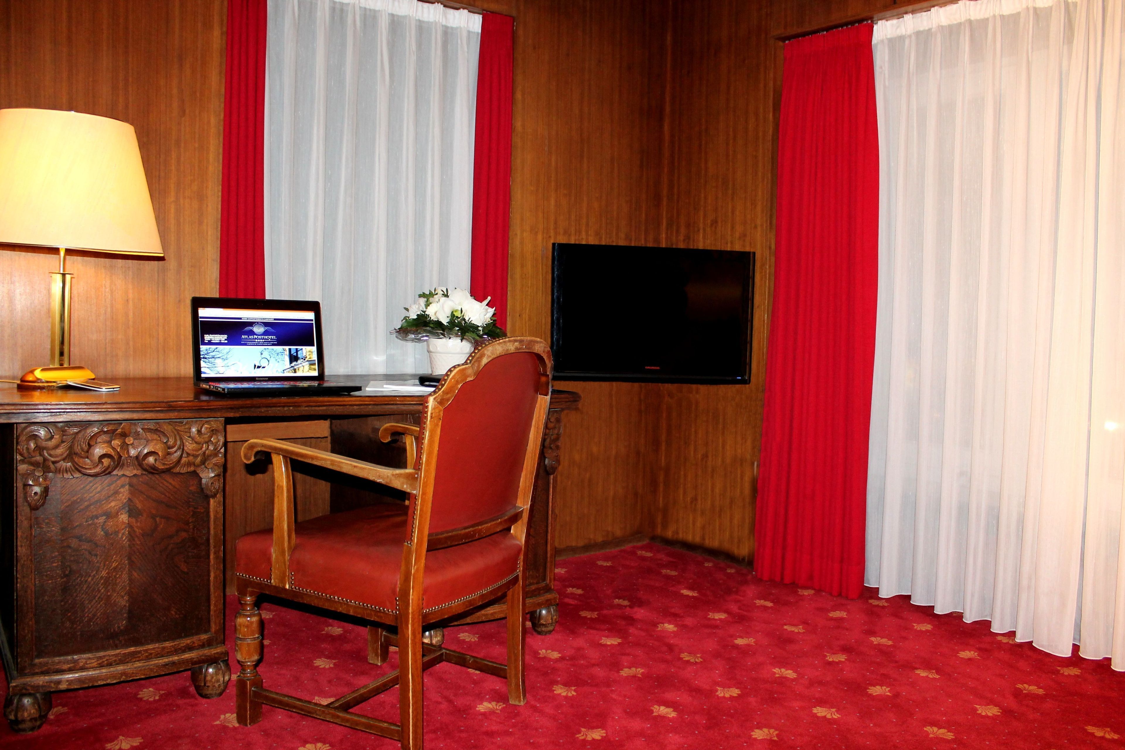A hotel room with a wooden desk, a laptop, a television, and a red chair.