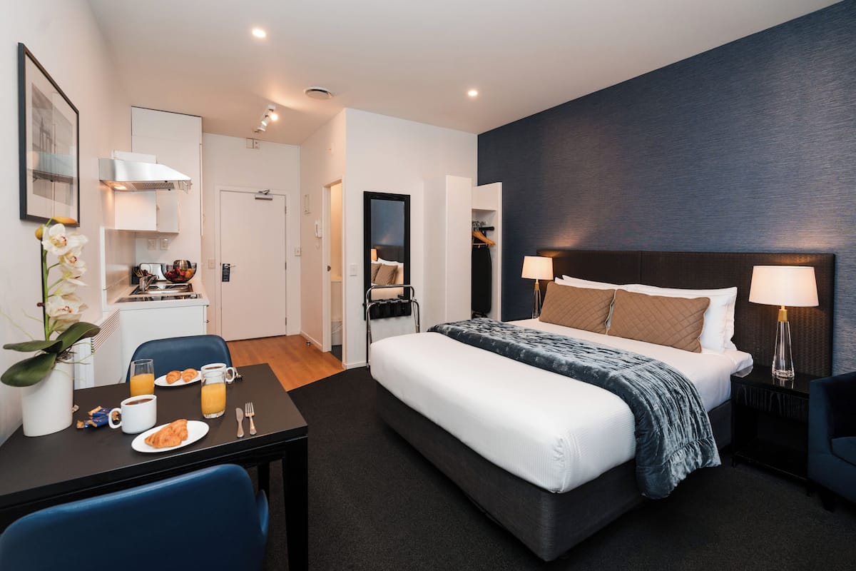 Una habitación de hotel moderna con una cama grande, mesitas de noche, un escritorio con desayuno y una kitchenette pequeña.