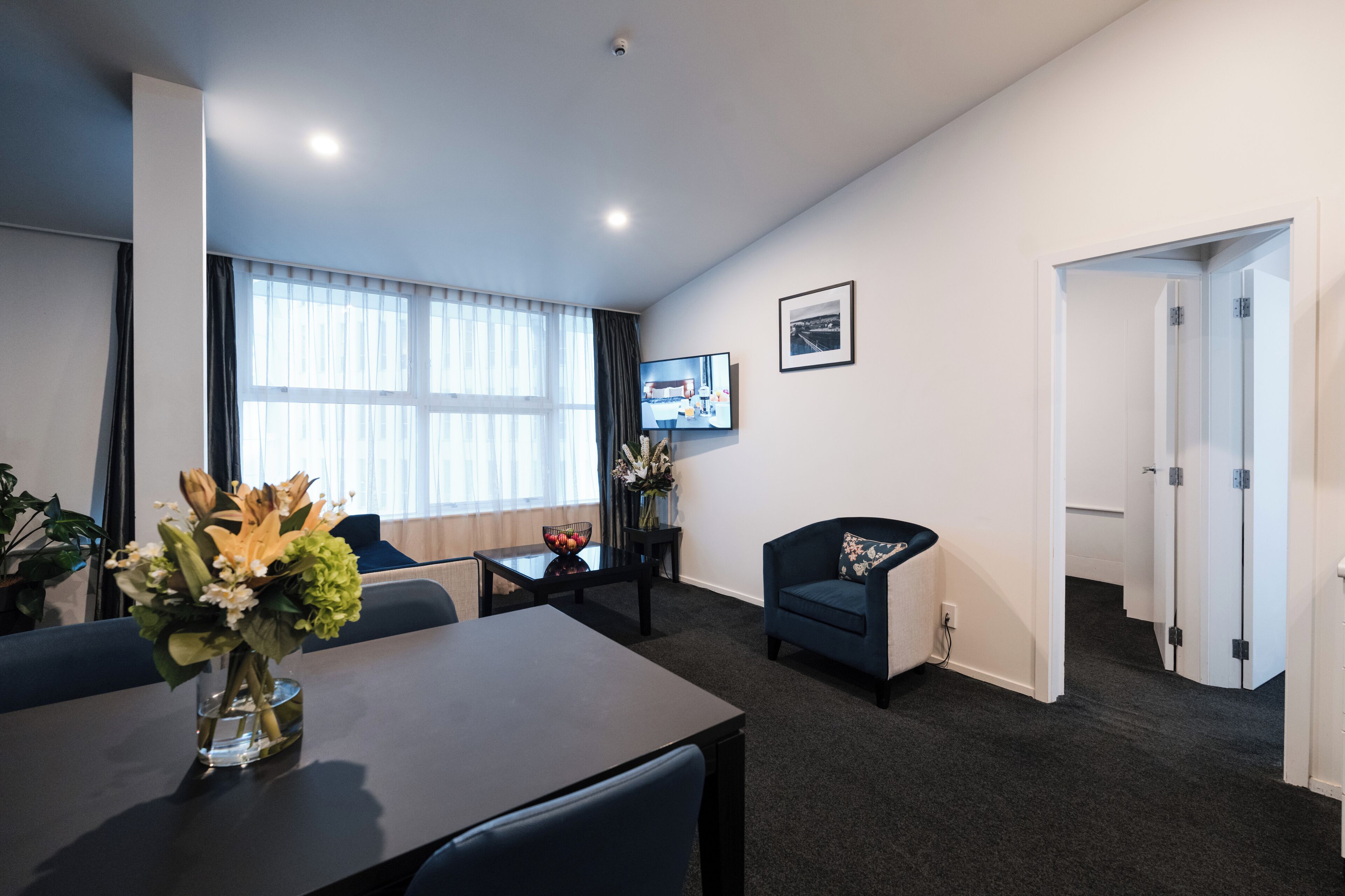 A modern hotel room with a large window, a flat-screen TV, a dining table with a vase of flowers, a blue armchair, and a door leading to another room.