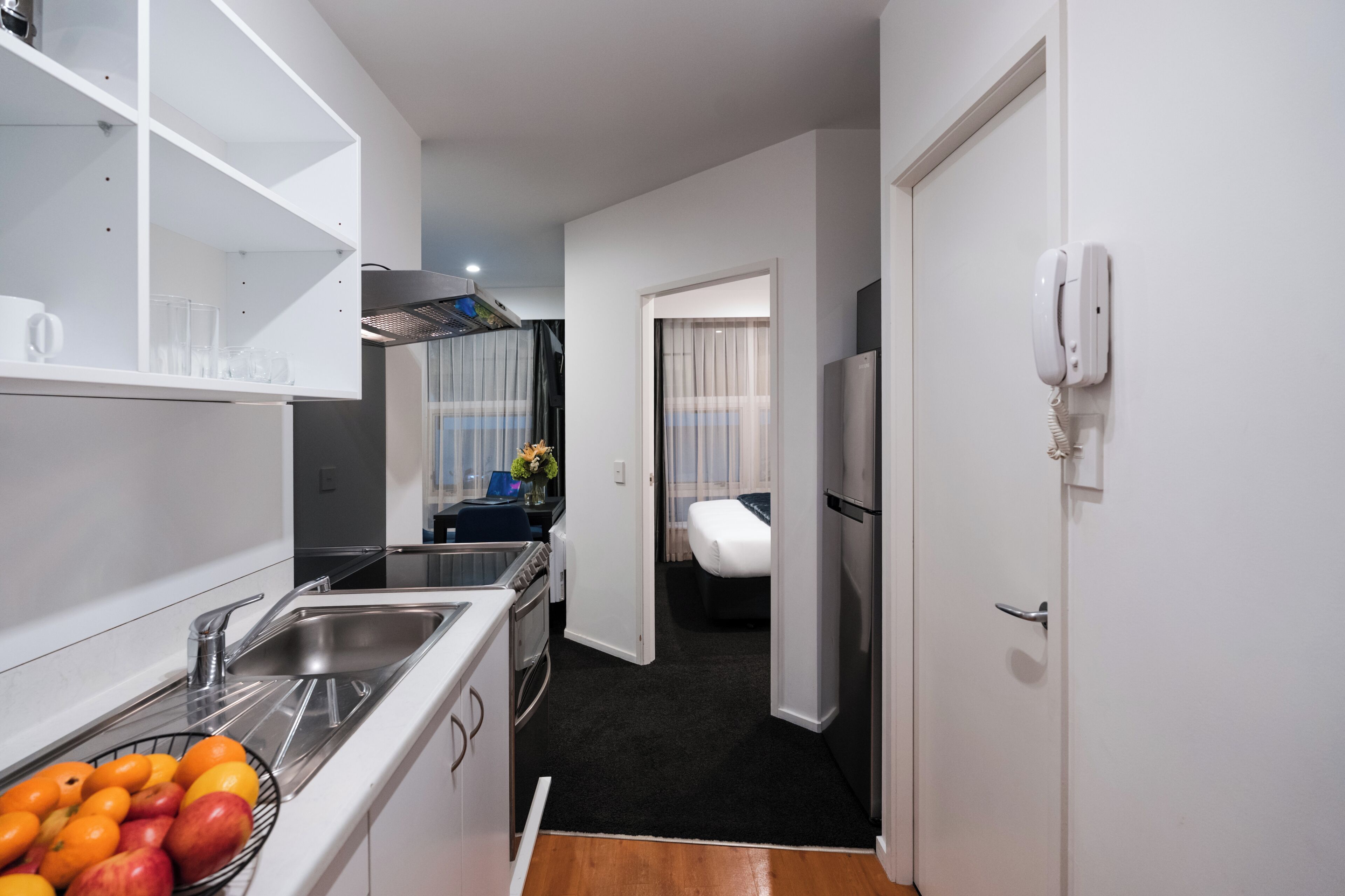 Une cuisine compacte équipée d’un évier, d’une cuisinière et d’un réfrigérateur. On y trouve un petit coin repas avec une chaise et une table. Un lit est visible à travers une porte ouverte au fond de la pièce.