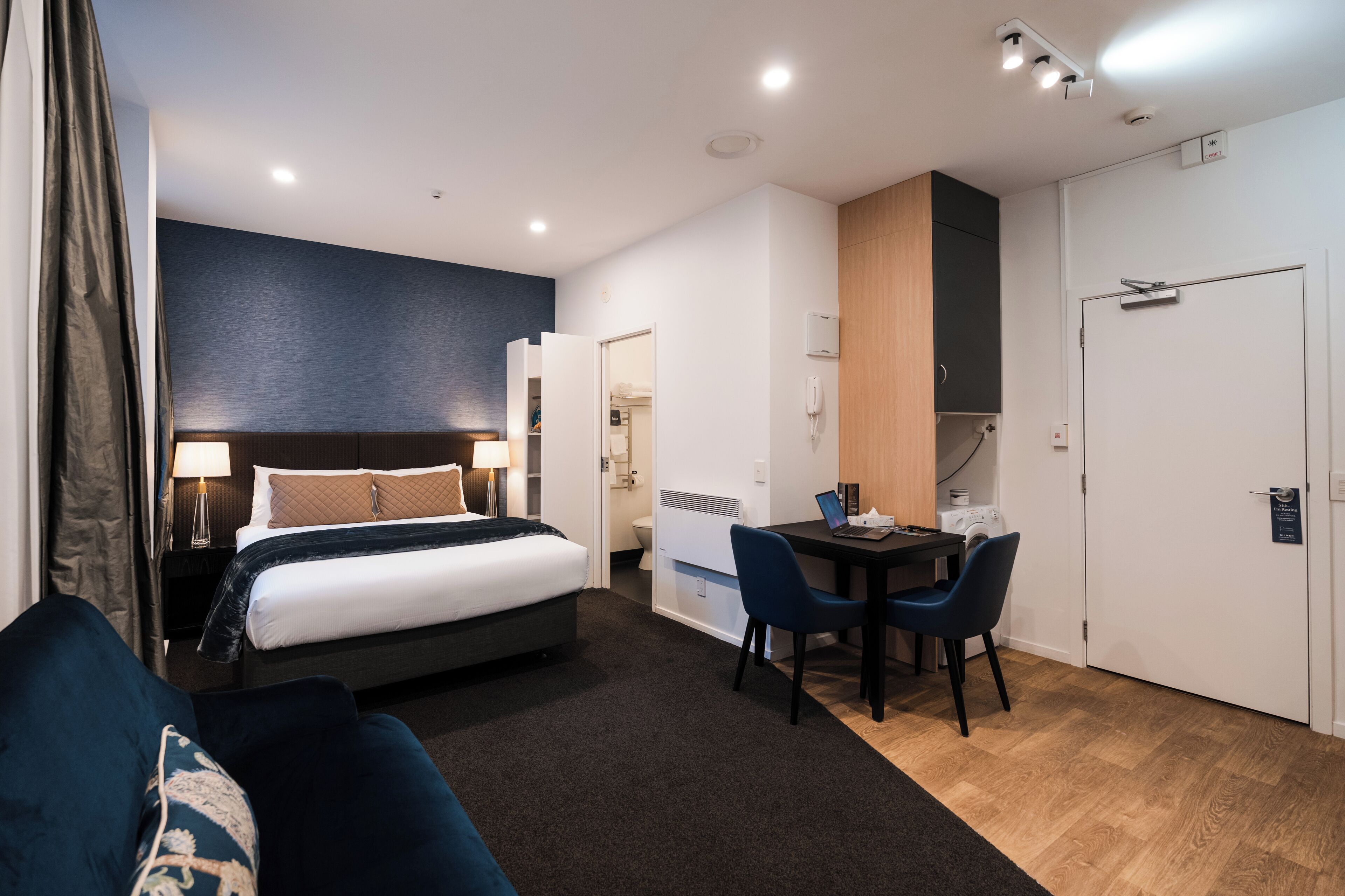 A modern hotel room with a bed, bedside table, lamp, and a small desk with a chair.