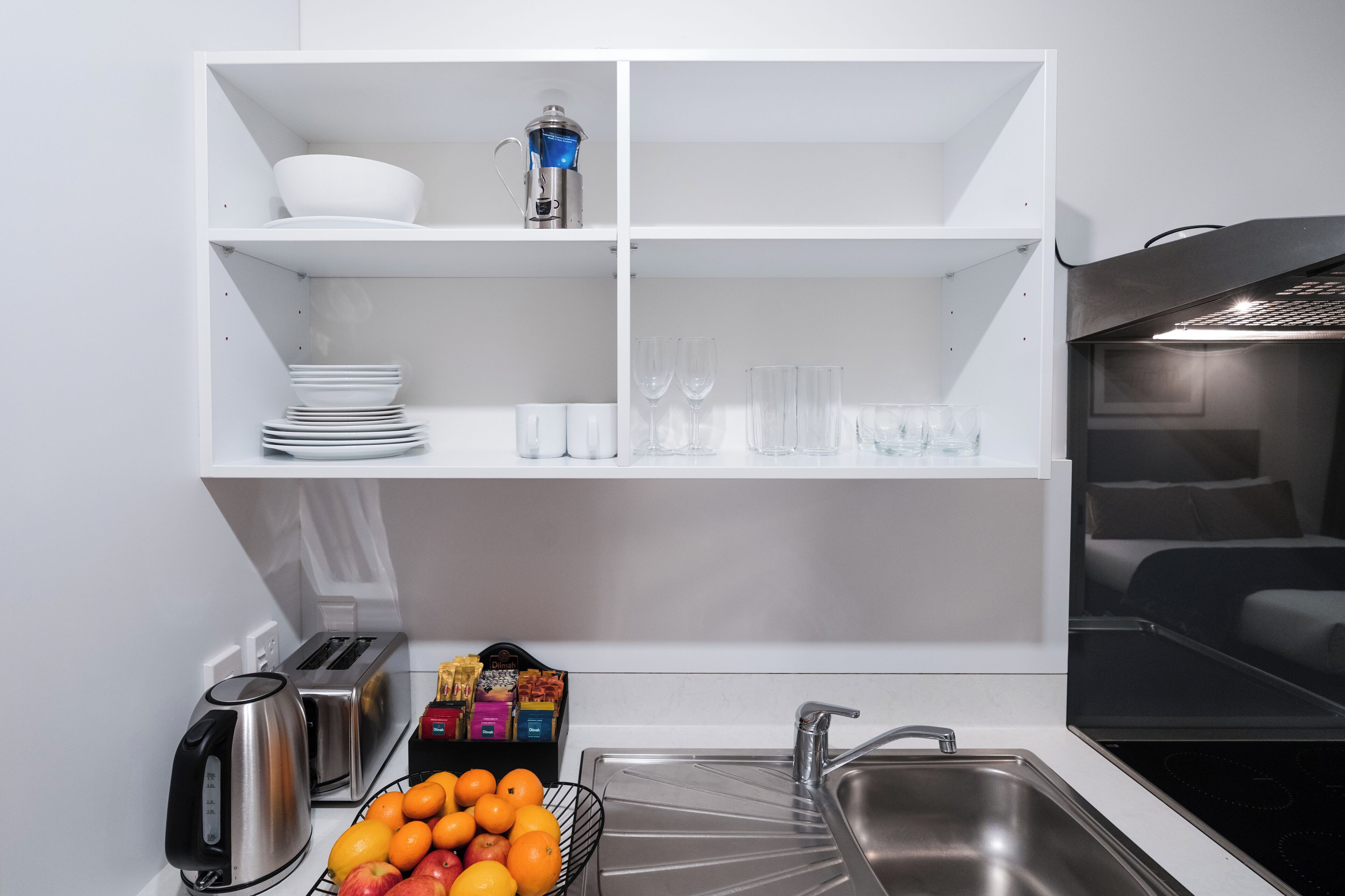 Een moderne keuken met witte kastjes, een inbouwfornuis en een spoelbak. Er zijn planken met servies en glazen, en een fruitmand met sinaasappels en appels.