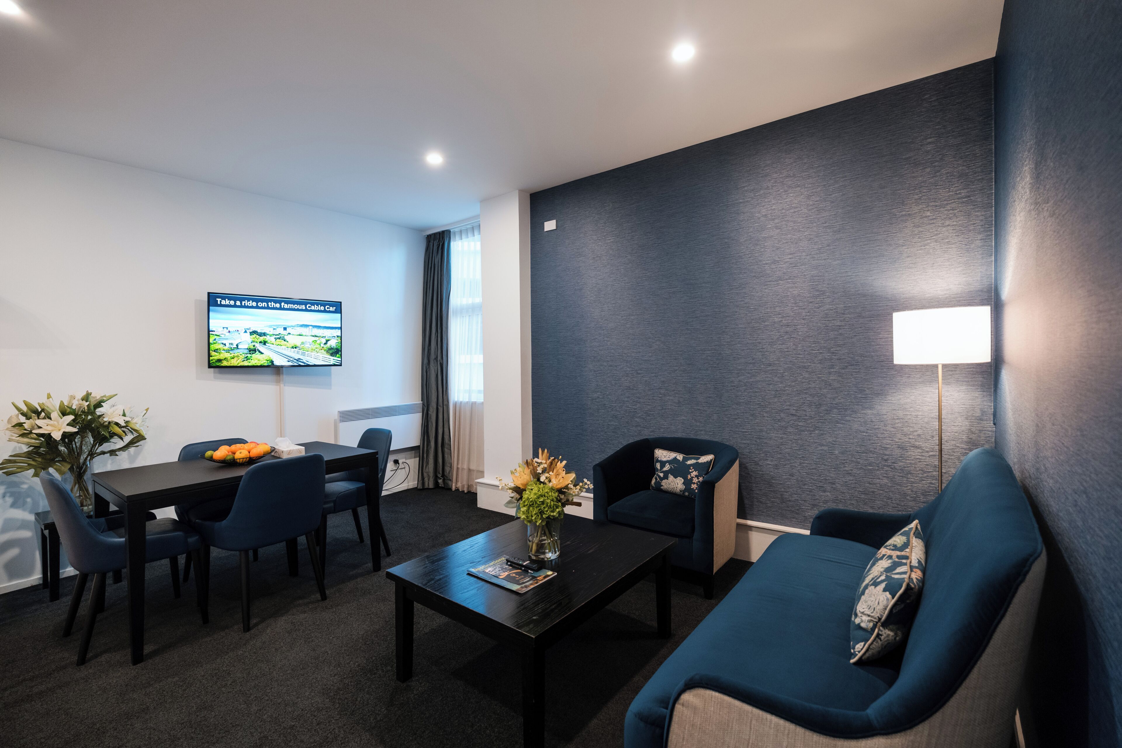 Una habitación de hotel moderna con un sofá azul oscuro, una mesa de comedor con sillas, un televisor de pantalla plana montado en la pared y un jarrón con flores sobre la mesa auxiliar.