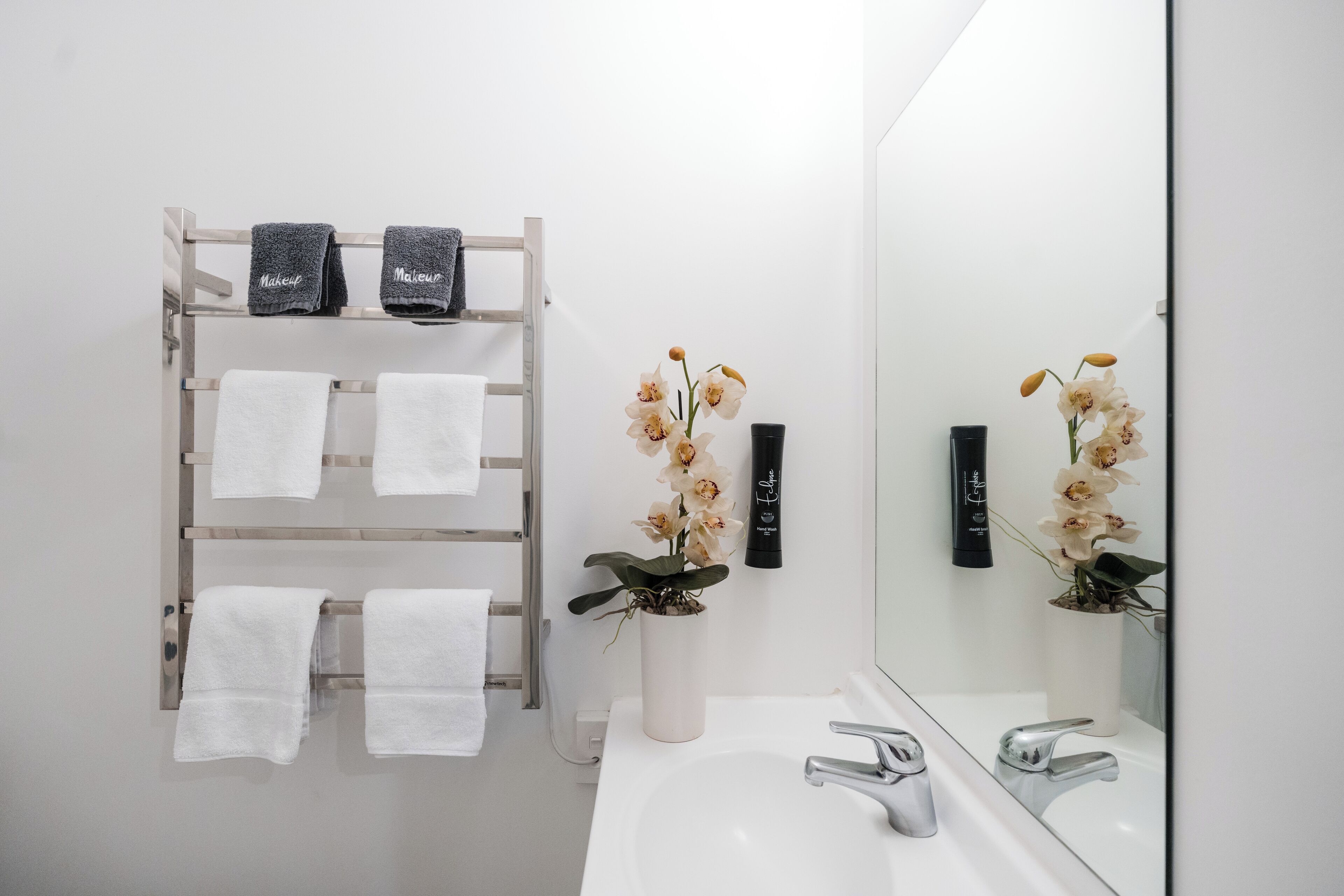 Un baño con lavamanos blanco, dos toalleros con toallas blancas y negras, y un florero con orquídeas.