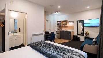 A modern hotel room with a bed, a desk, a chair, a TV, and a kitchenette.