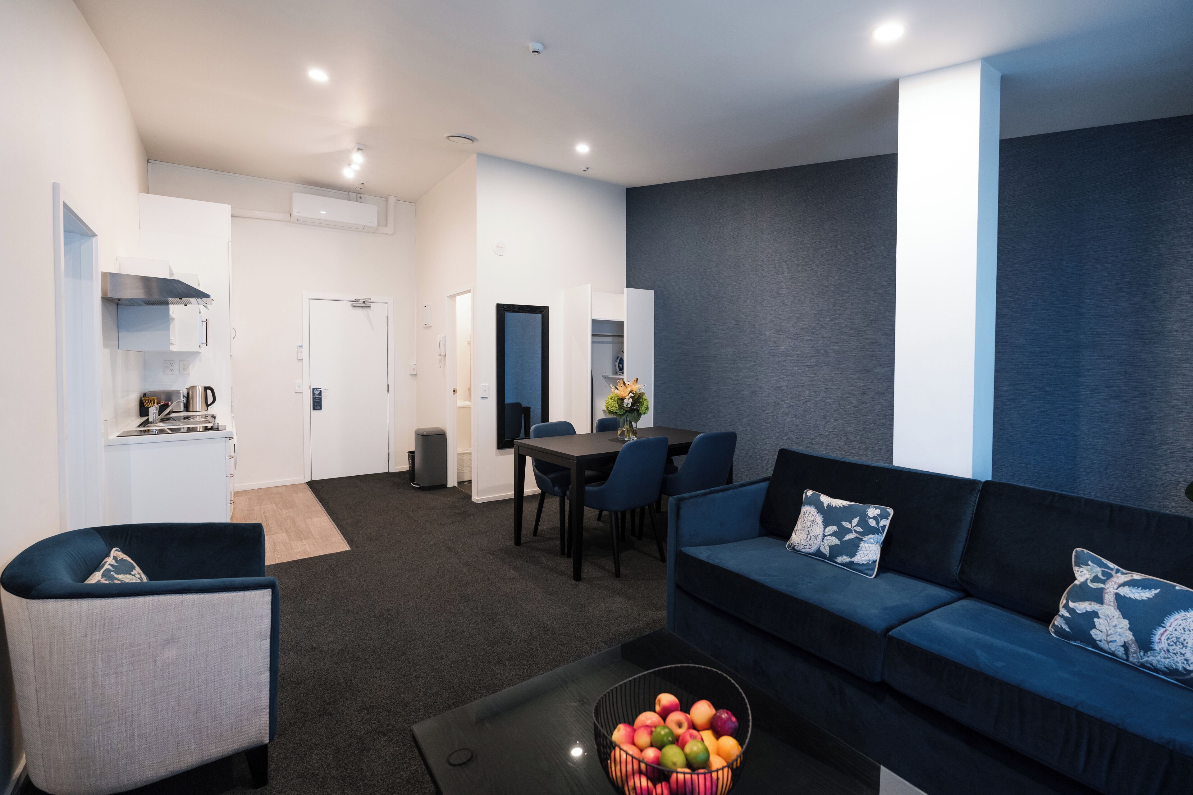 A modern living room with a dark blue sofa, a small armchair, a dining table with chairs, and a kitchen area with a stove and cabinets.