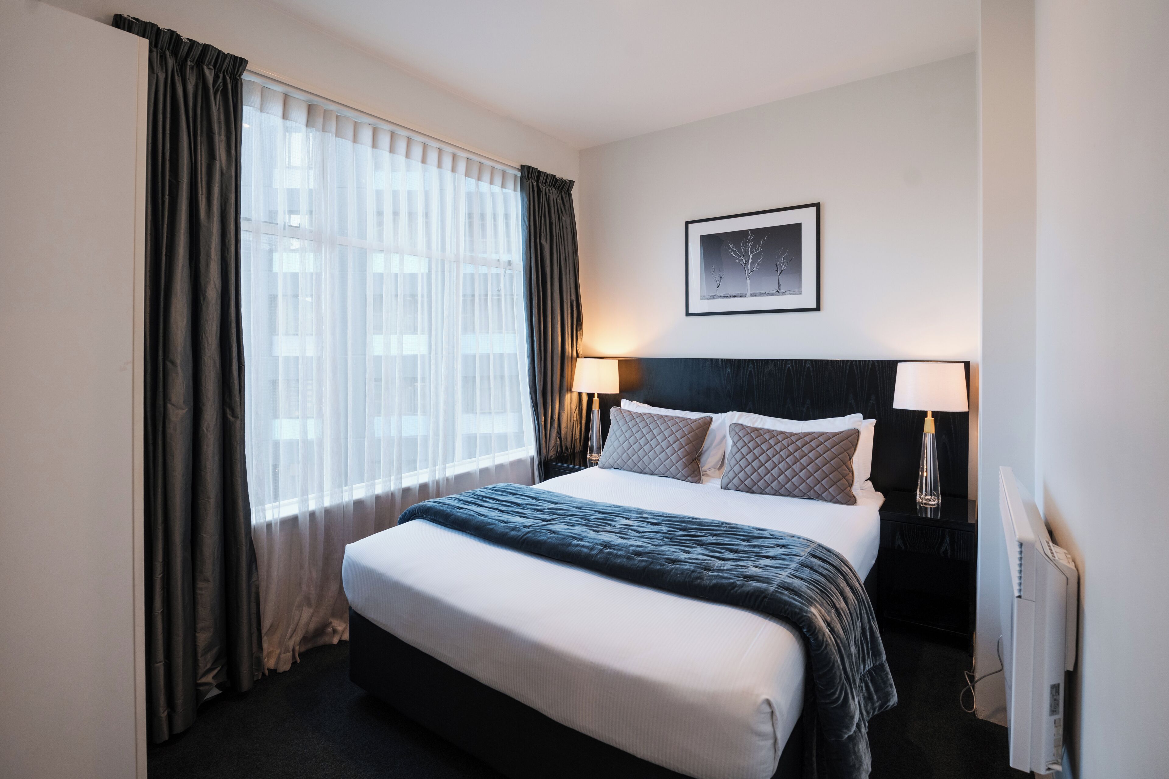 A hotel room with a large bed, two bedside lamps, a framed picture on the wall, and a window with sheer curtains.