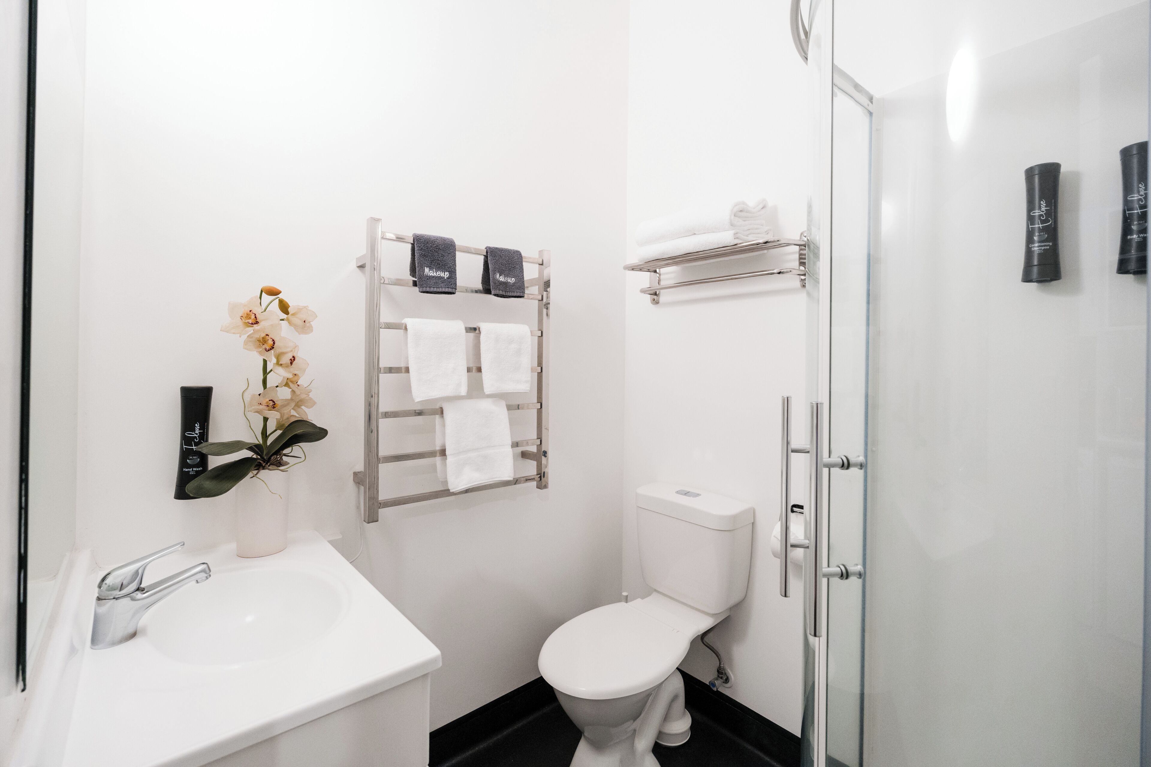 A modern bathroom with a white sink, toilet, and shower. There are towels and toiletries on shelves.