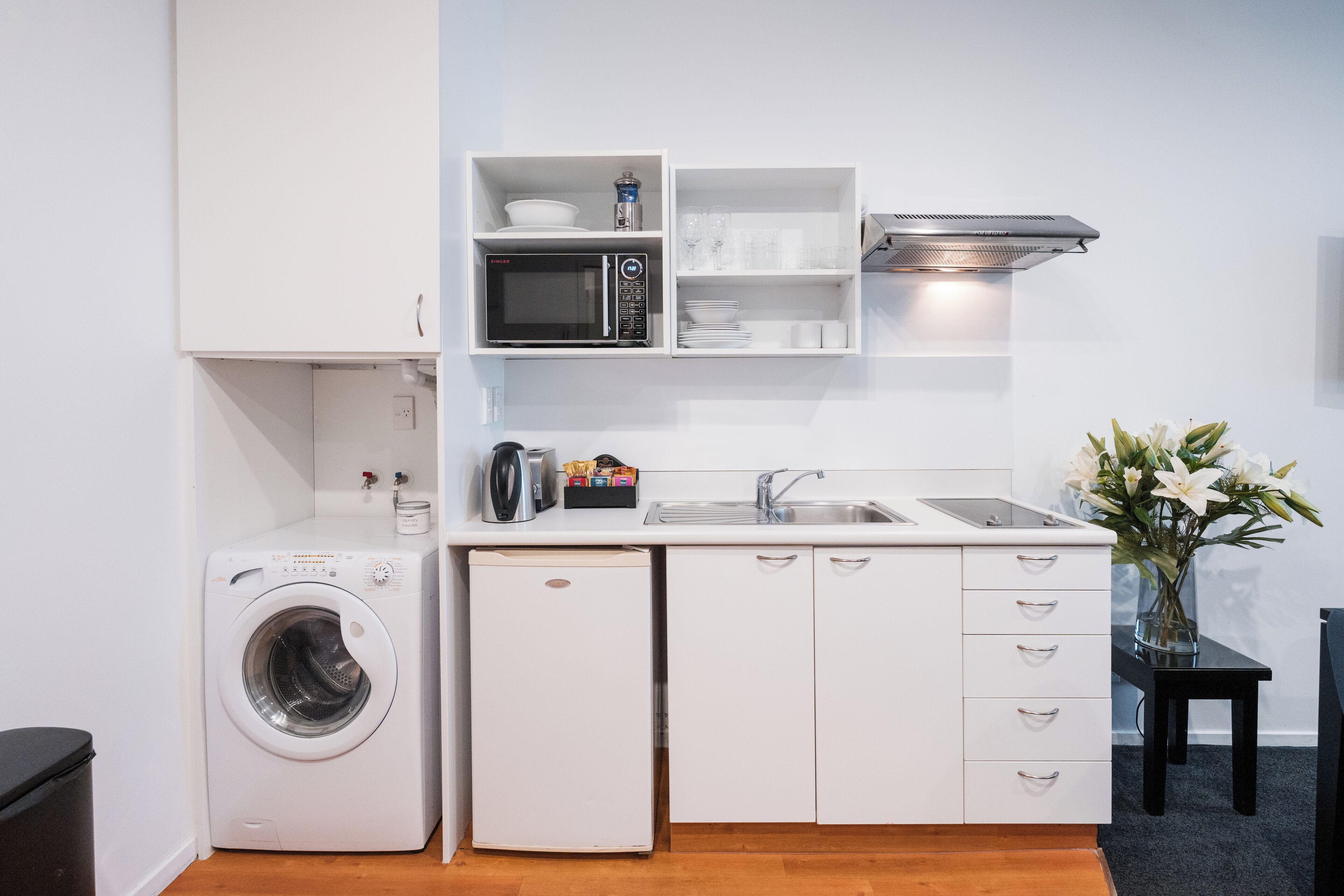 A compact kitchen with a washing machine, a small refrigerator, white cabinetry, a microwave, and a sink.