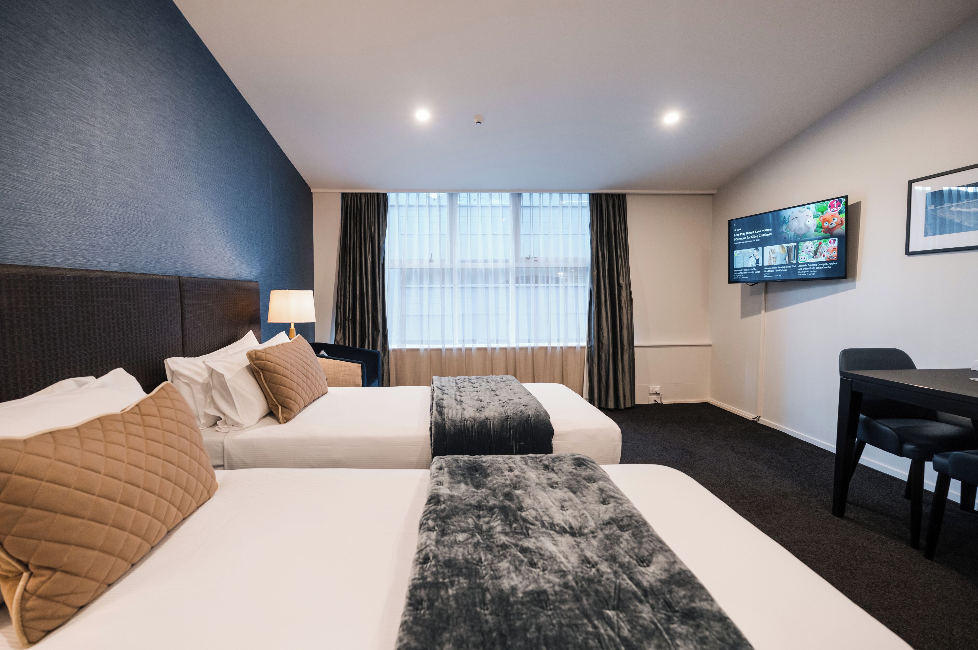 A hotel room with two beds, a desk, a TV, and a window with curtains.