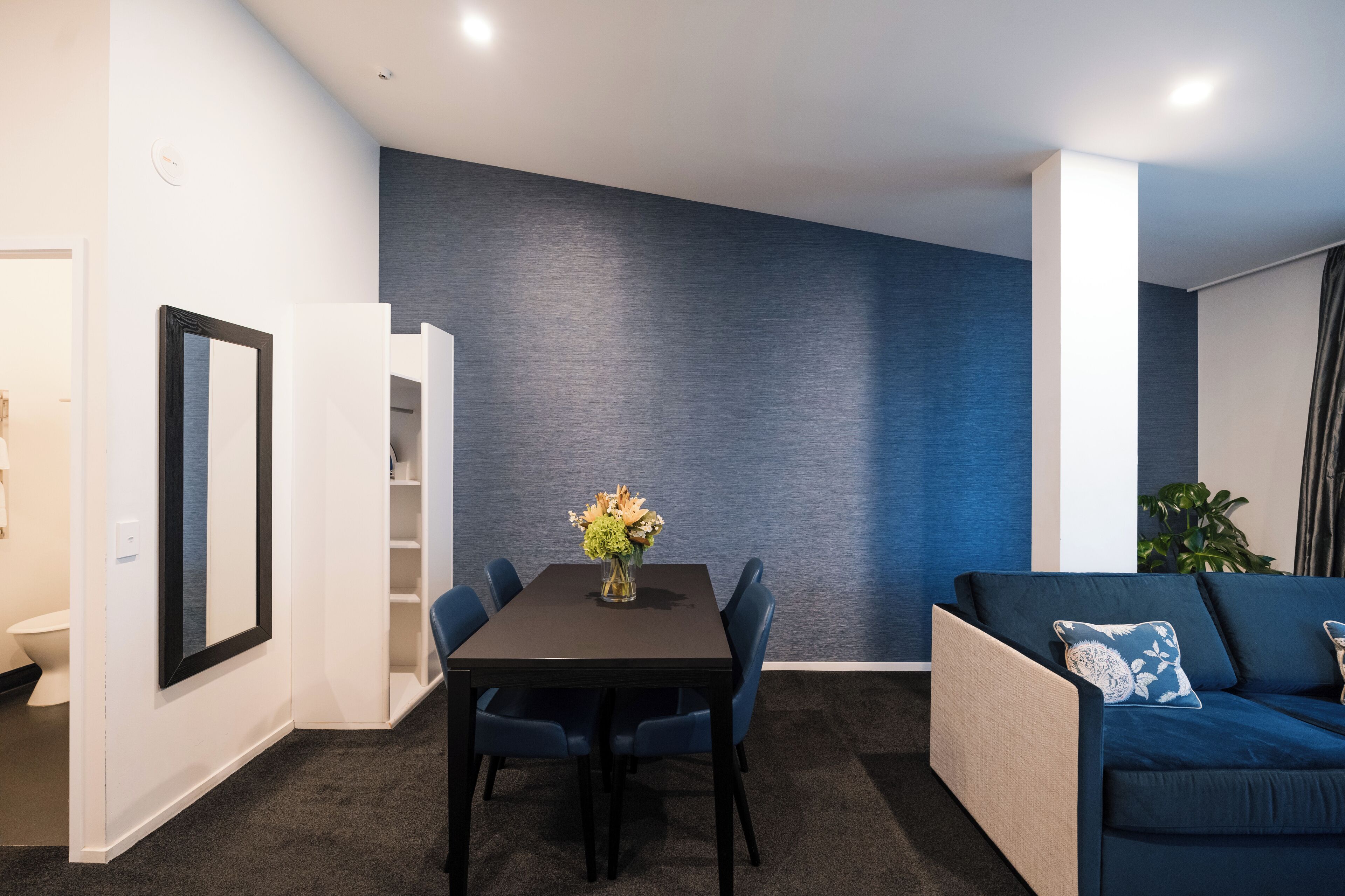 A modern room with a dark blue accent wall, a dark wooden table with blue chairs, a blue sofa with decorative pillows, and a small plant.