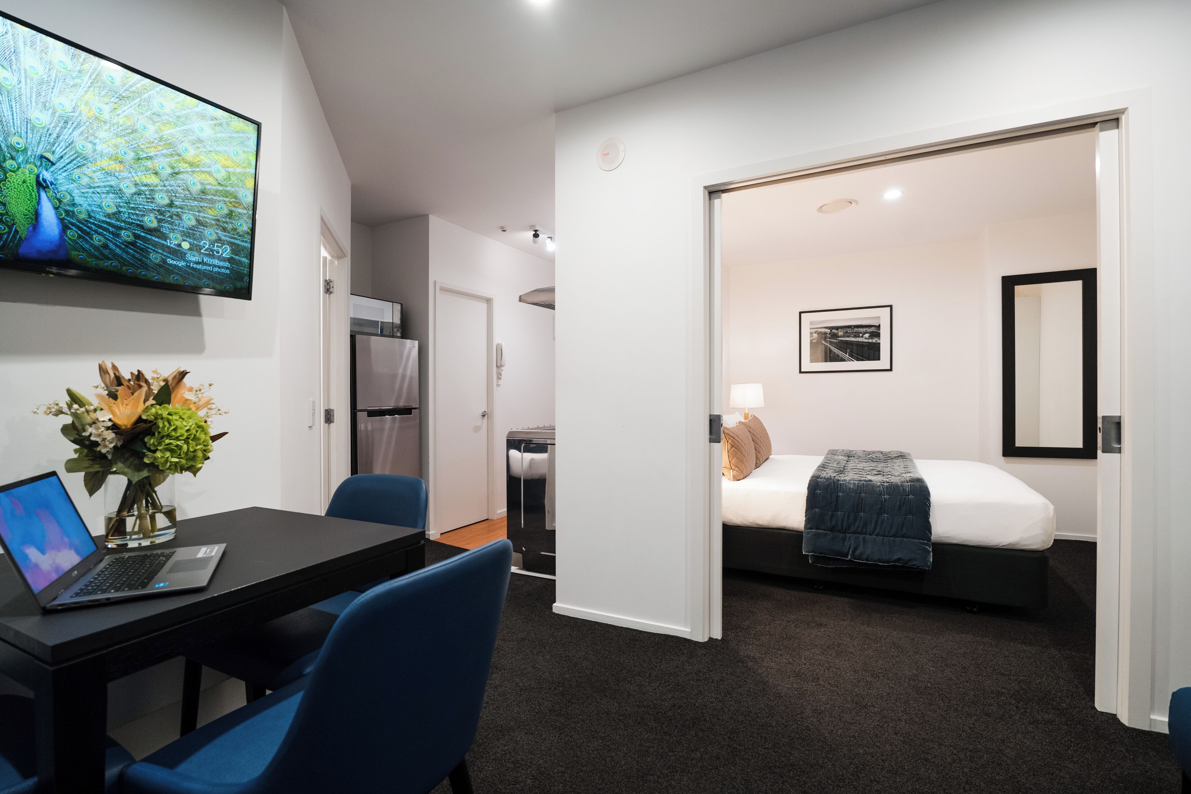 Ein modernes Hotelzimmer mit einem Bett, einem Schreibtisch mit einem Laptop, einem an der Wand montierten Fernseher und einem durchgehenden Türspalt einsehbaren Badezimmer.