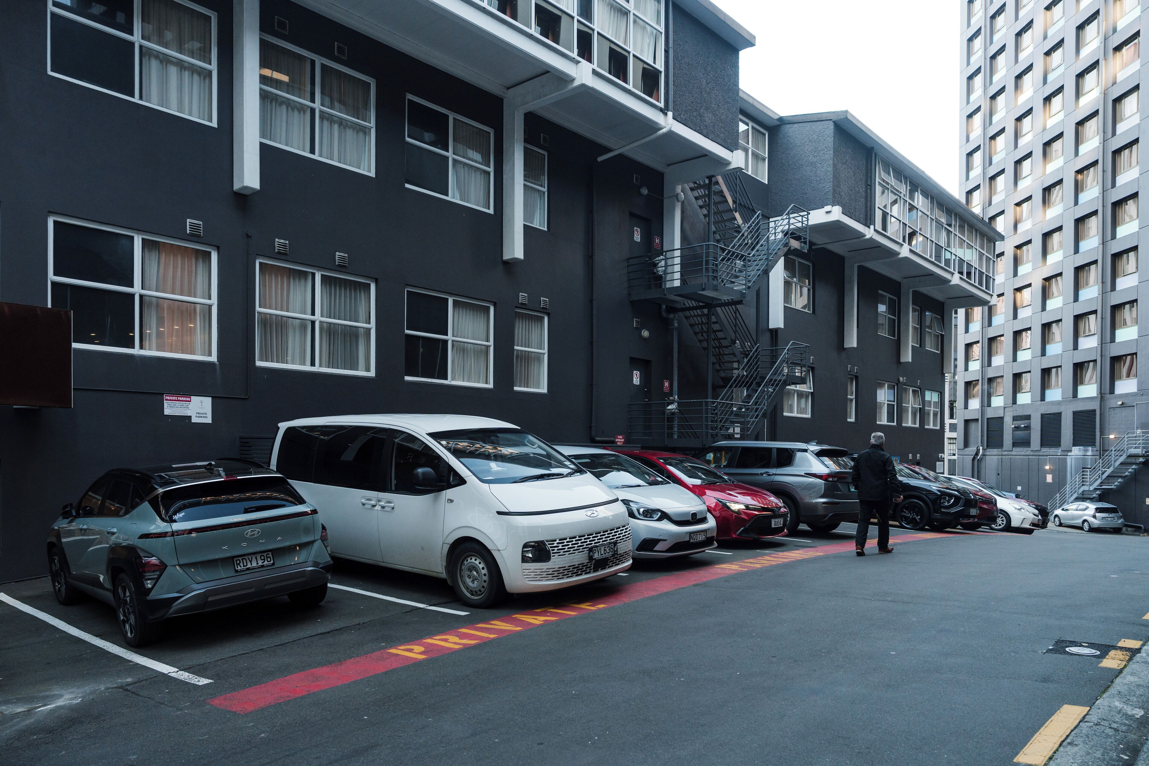 A parking lot with several cars, including a white van and a red car, parked in front of a multi-story building with a fire escape.