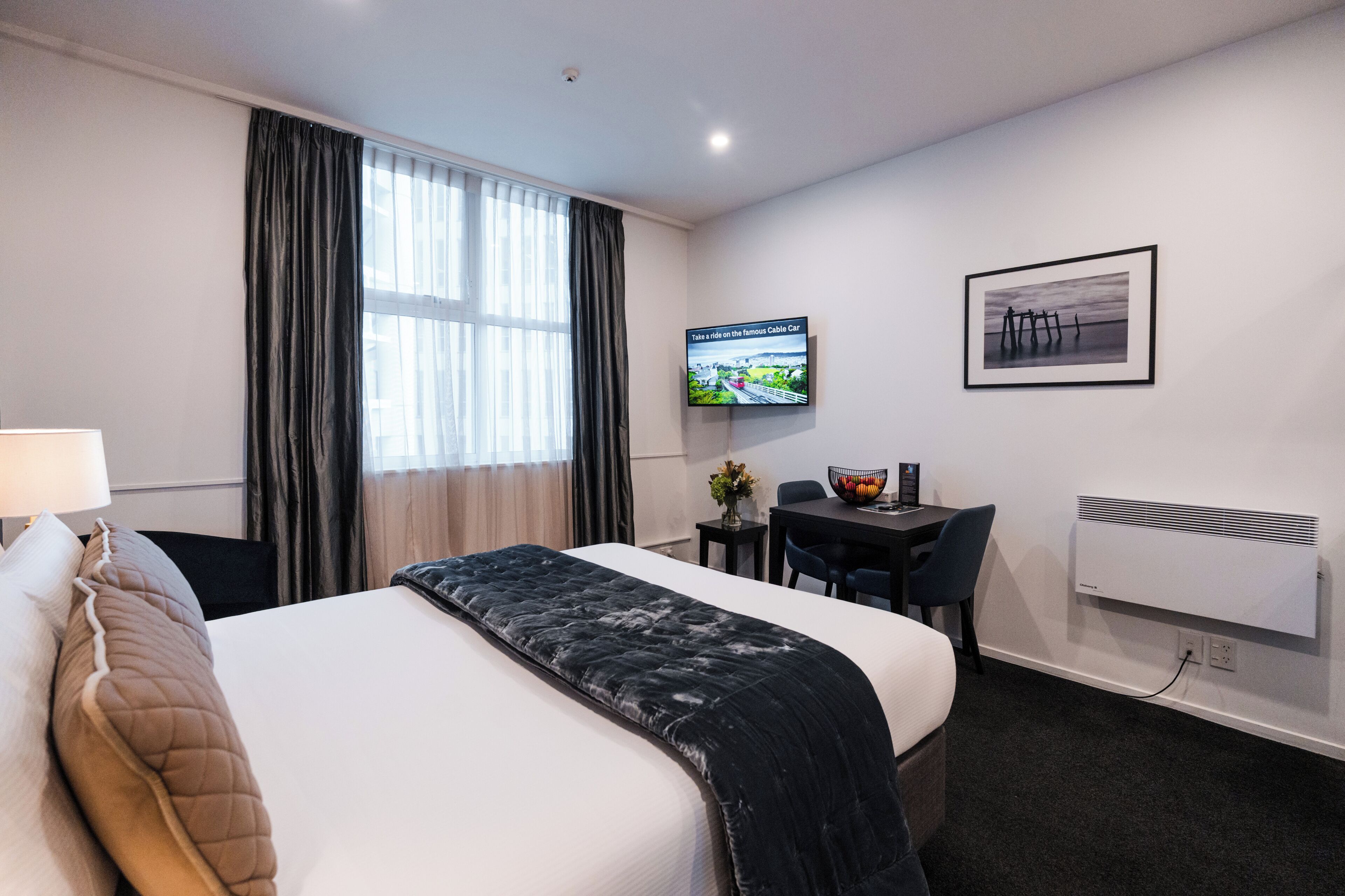 A hotel room with a bed, a desk, a chair, a TV, and a radiator.