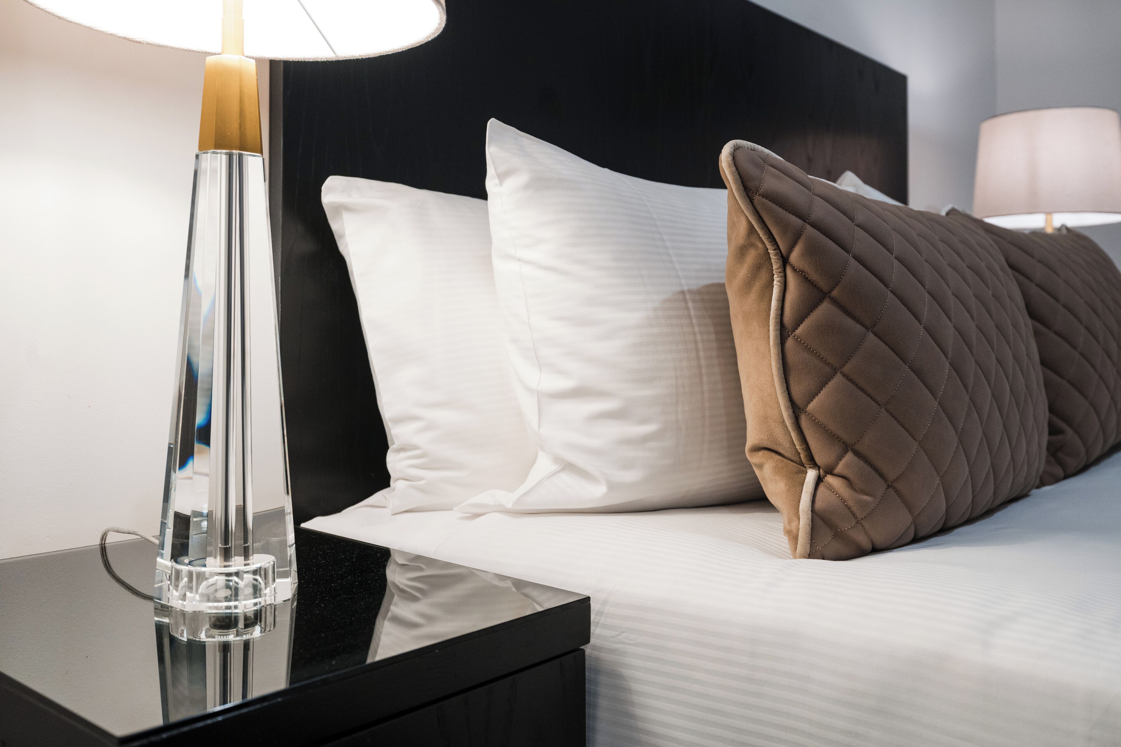 Cama bem arrumada com travesseiros brancos e castanhos, um vaso de vidro transparente sobre uma mesa de cabeceira preta, e uma luminária na mesa de cabeceira.