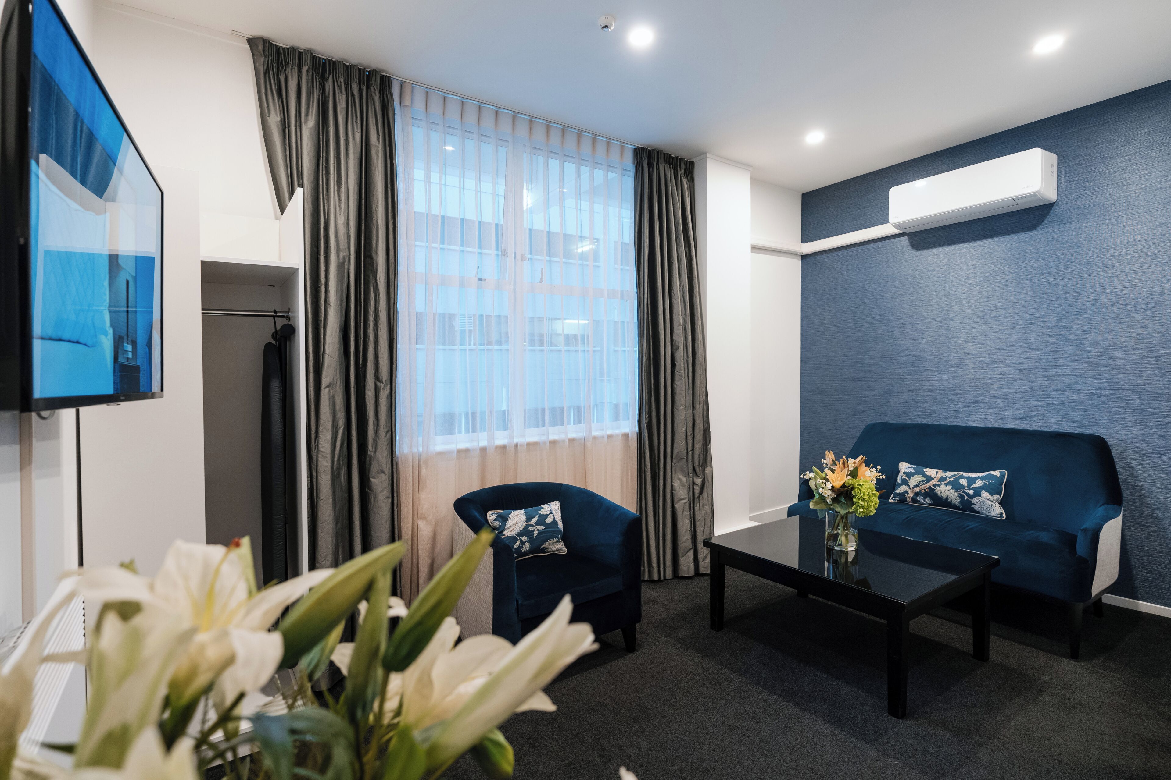 Una habitación de hotel moderna con un sofá azul oscuro, una mesita, un televisor y un ventanal con cortinas.