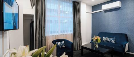 Una habitación de hotel moderna con un sofá azul oscuro, una mesita, un televisor y un ventanal con cortinas.